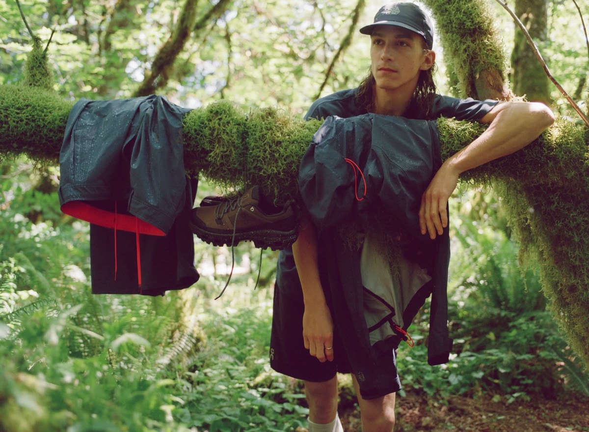 A person leans on a moss-covered branch, on which are draped a few pieces of outdoor gear including rainwear and a pair of boots.