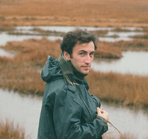 A young man with brown hair standing in front of a marsh, wearing an olive-colored rain jacket. His head is turned towards the camera.