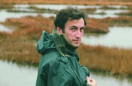 A young man with brown hair standing in front of a marsh, wearing an olive-colored rain jacket. His head is turned towards the camera.
