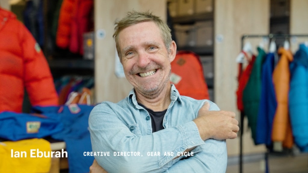 A person smiles at the camera in front of garments in a storeroom. Underneath his picture is a line of text that reads Ian Eburah, creative director, gear and cycle