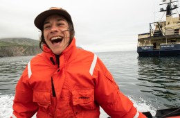 A person wearing a bright orange jacket on a raft in the water laughs with an open mouth.