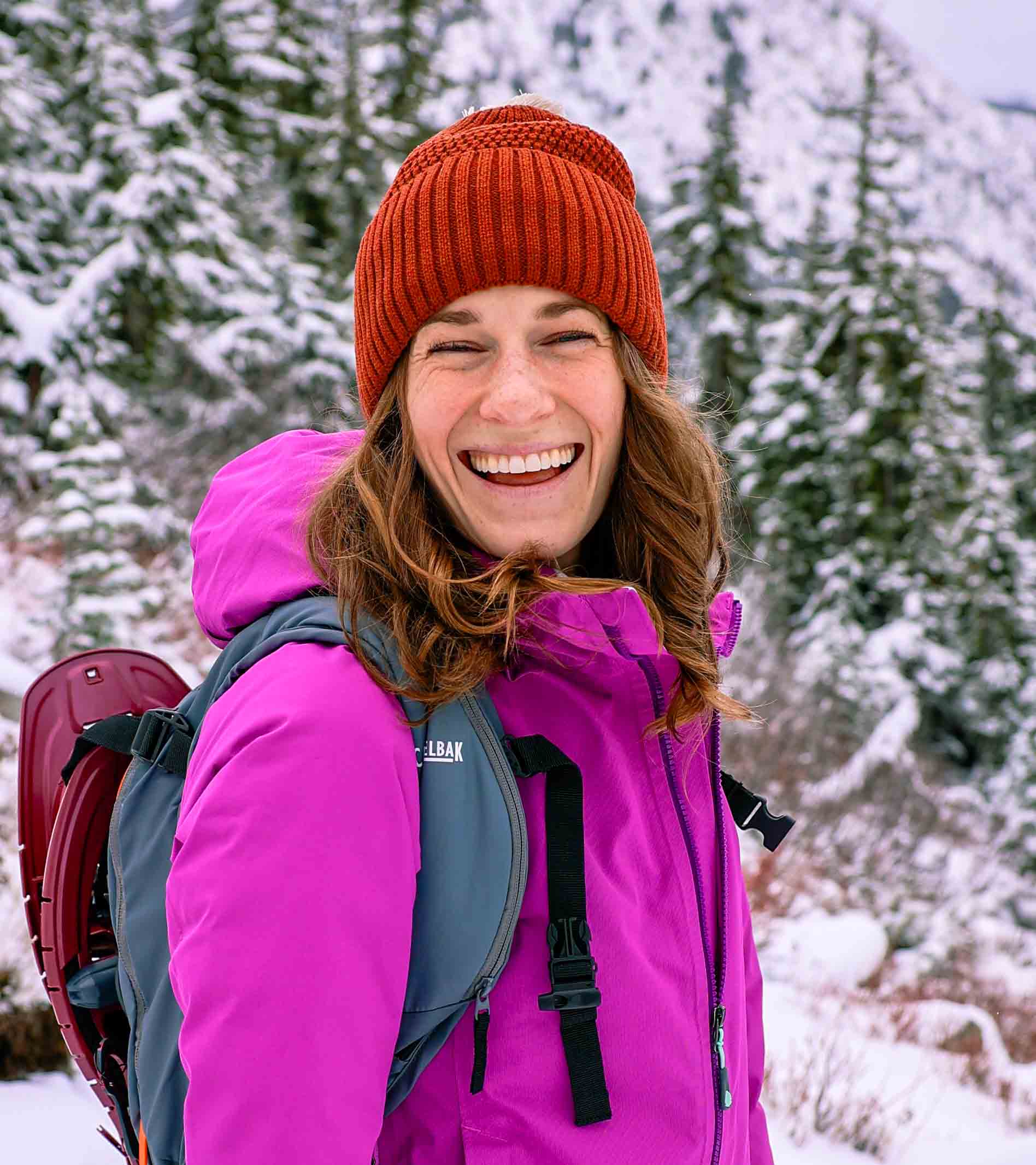 A person wearing a magenta jacket and brick-red beanie laughs in a snowy woods.