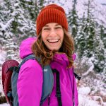 A person wearing a magenta jacket and brick-red beanie laughs in a snowy woods.
