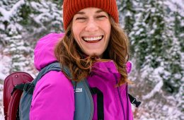 A person wearing a magenta jacket and brick-red beanie laughs in a snowy woods.
