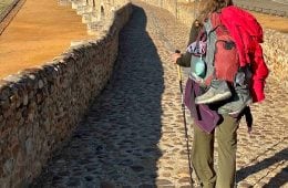 A shot from behind of a hiker carrying a trekking pole and wearing a loaded down pack on a long stone arched bridge.