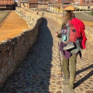 A shot from behind of a hiker carrying a trekking pole and wearing a loaded down pack on a long stone arched bridge.
