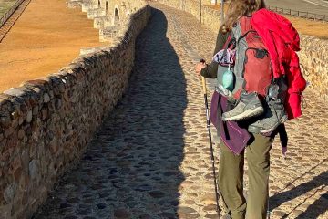 A shot from behind of a hiker carrying a trekking pole and wearing a loaded down pack on a long stone arched bridge.