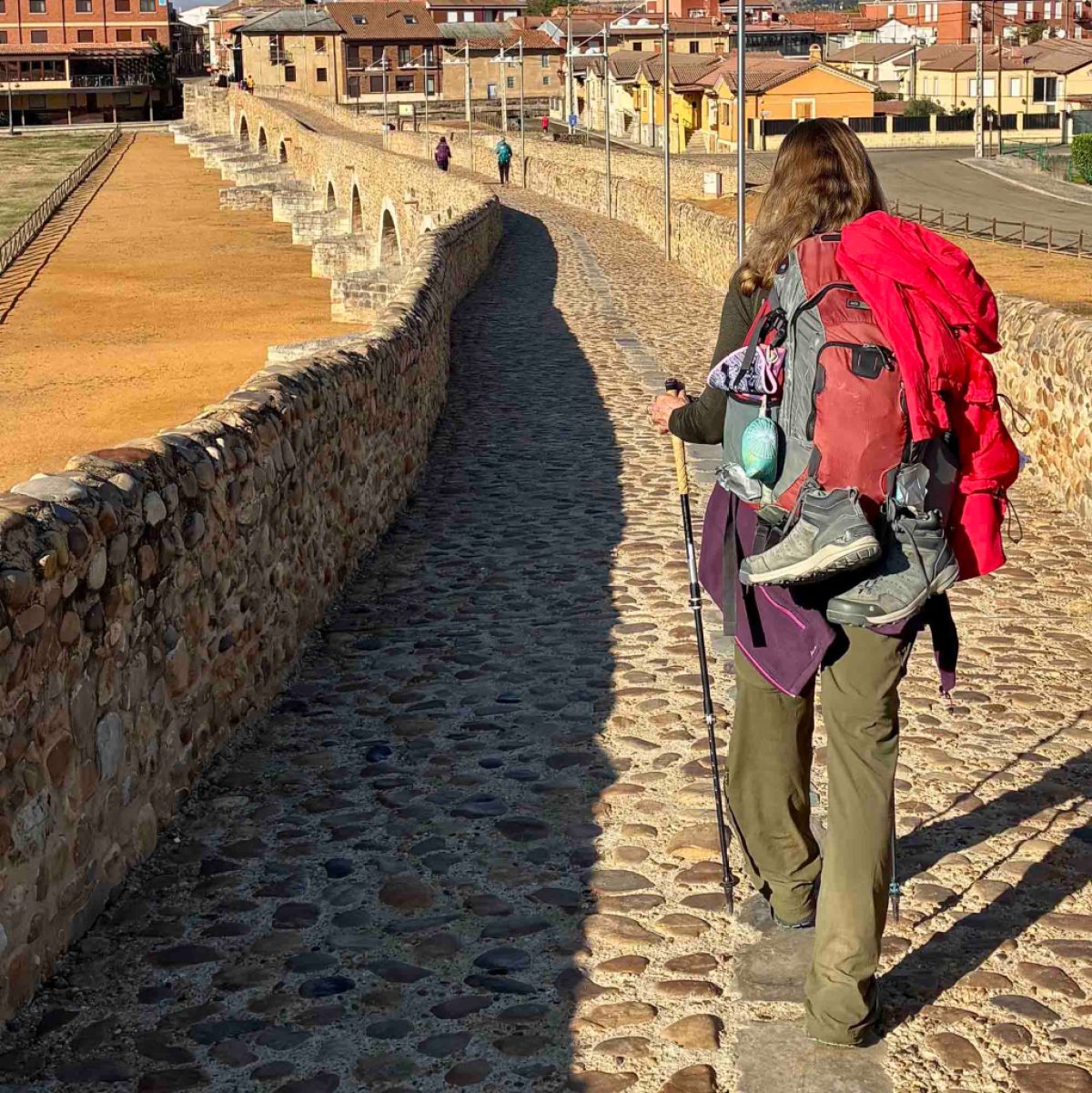 A shot from behind of a hiker carrying a trekking pole and wearing a loaded down pack on a long stone arched bridge.