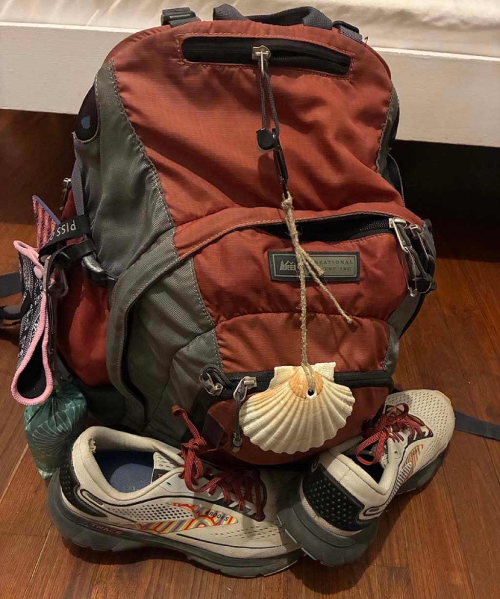 A used red backpack with a seashell tied to it and some hiking shoes sits on the floor of a room.