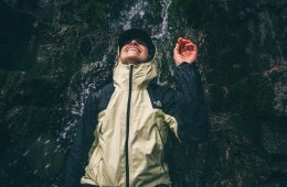 A person standing under a waterfall wearing a hooded rain jacket smiles.