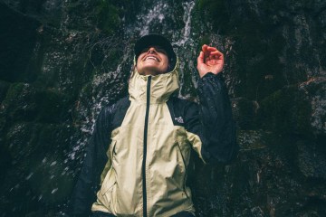 A person standing under a waterfall wearing a hooded rain jacket smiles.