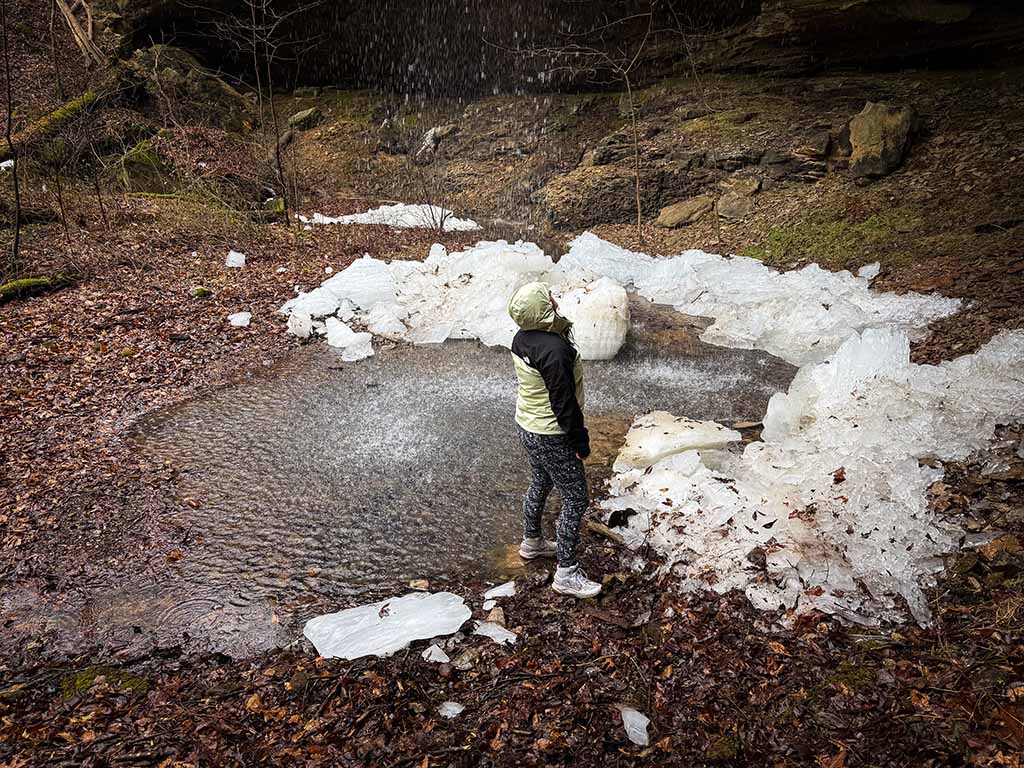 A person wearing a black and olive colored rain jacket stands with their back turned to the camera, standing under a waterfall. 
