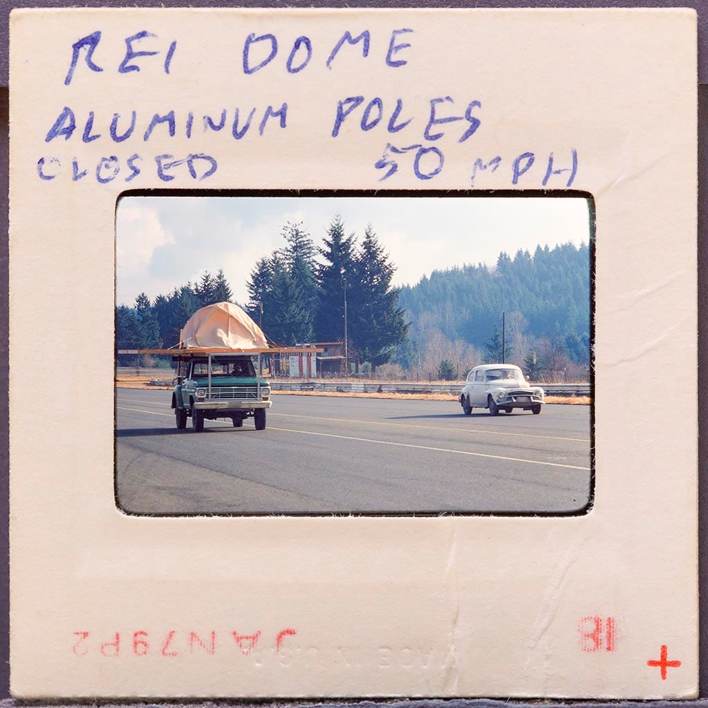 An old photo of a tent strapped to a truck riding down the highway. On the photo is written "REI Dome Aluminum Poles. Closed 50 mph."