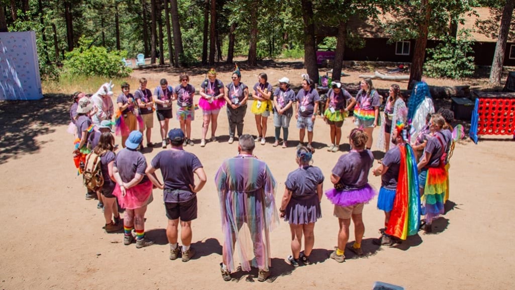 A group of LGBTQ+ youth stand in a circle at a campground, many wearing festive or rainbow outfits.