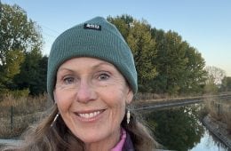 A person wearing a blue beanie and a red fleece smiling at the camera under a twilight sky.