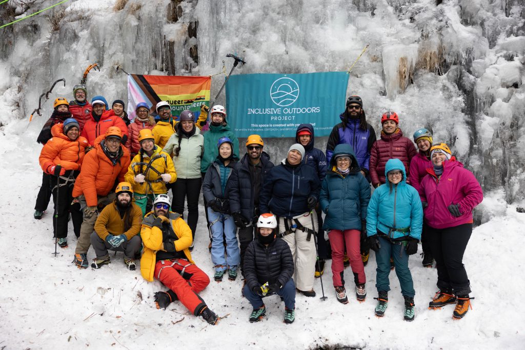 A group of people smiling on an icy mountain, holding up a banner that says Inclusive Outdoors Project as well as a rainbow progress Pride flag