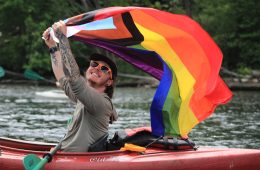 A person in a kayak flies a rainbow progress Pride flag over their head.