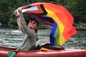 A person in a kayak flies a rainbow progress Pride flag over their head.