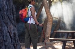 A person wearing hiking apparel, a hat and colorful backpack stands in front of a picnic table, holding trekking poles.
