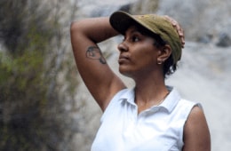 A person wearing a hat and a white sleeveless shirt standing in front of a rock face, with their hand on their head