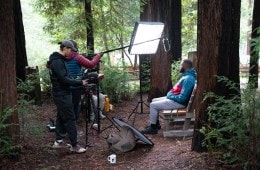Two people stand with camera, microphone and lighting equipment filming a black man sitting in front of redwood trees