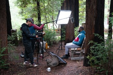 Two people stand with camera, microphone and lighting equipment filming a black man sitting in front of redwood trees