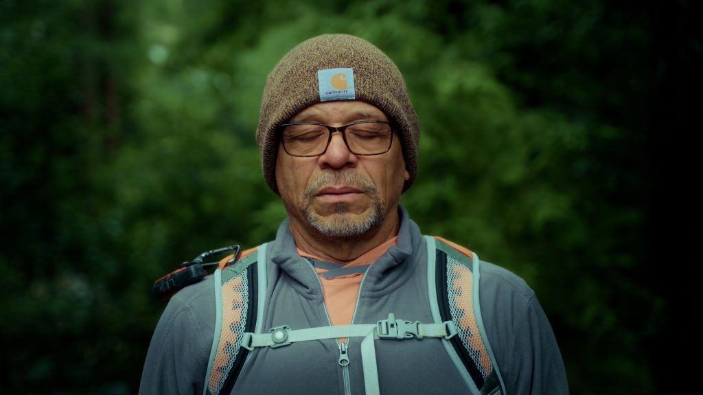 An older Black man with closed eyes wearing a backpack stands in the forest