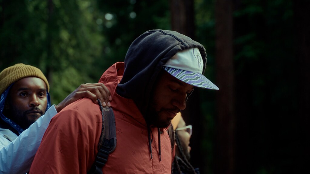 Two Black men standing in the woods, one with his hands on the other man's shoulders as he looks away.