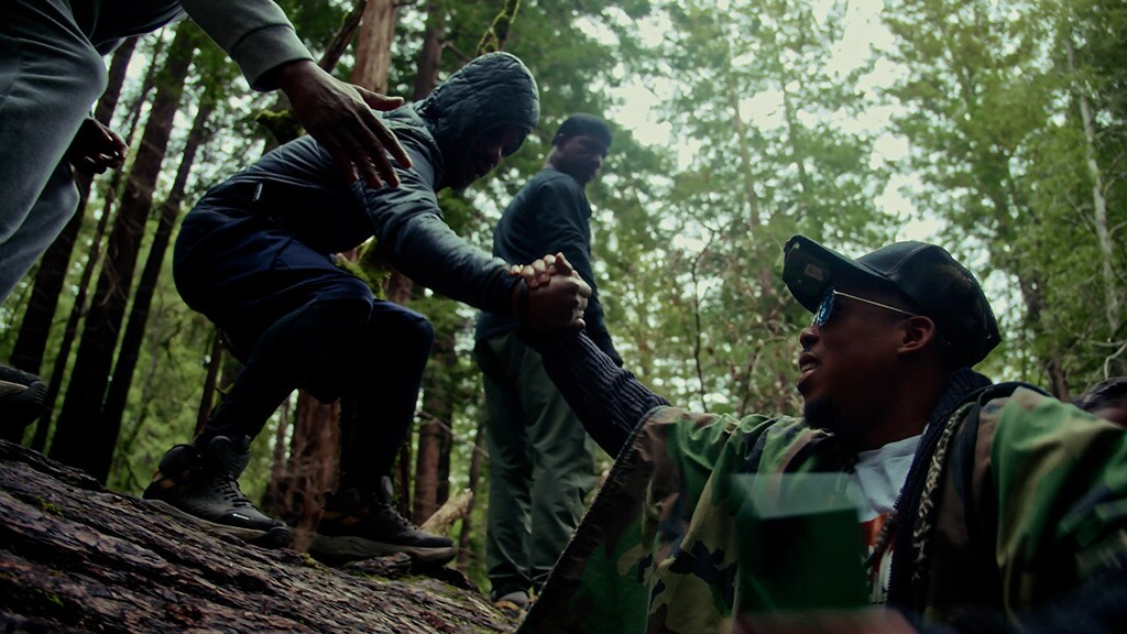 Two men standing on a high log reach down and help another climb up.