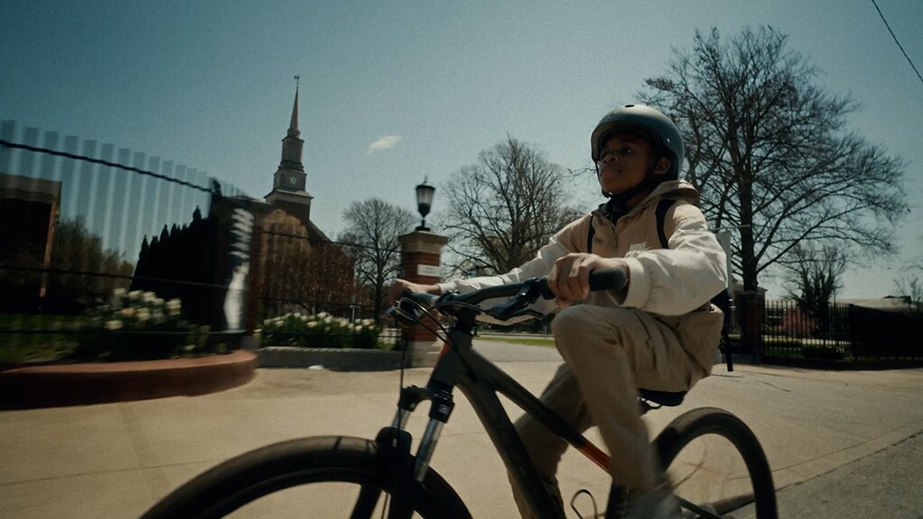 A young Black boy rides his bicycle in front of some large buildings and other structures in an urban environment
