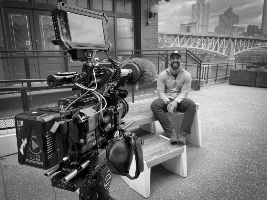 A black and white photo of a film camera aimed at a smiling man sitting on a bench