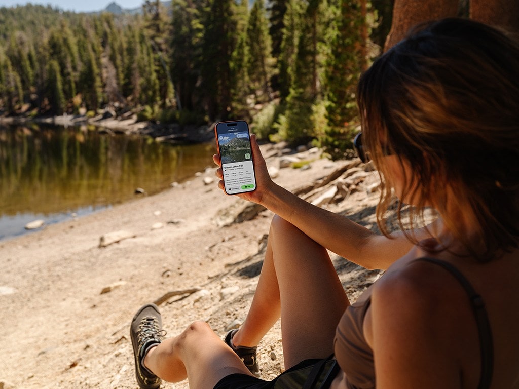 A person sitting up on the ground outdoors looks at their phone, which is displaying the app AllTrails