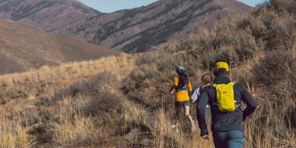 A man in a yellow hat, black jacket, and blue pants running behind two women in a mountain valley. He is wearing the REI Swiftland 5 Hydration Vest in yellow.