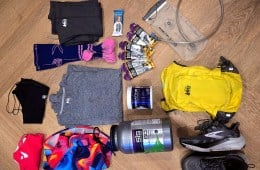 Author Rebecca Campbell's ultramarthon training kit, including a yellow REI Co-op hydration vest, black Brooks Ghost trail running shoes, a gray shirt, black running tights, multi-color running shorts, running gels, gaiters, and hydration powder.