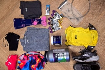 Author Rebecca Campbell's ultramarthon training kit, including a yellow REI Co-op hydration vest, black Brooks Ghost trail running shoes, a gray shirt, black running tights, multi-color running shorts, running gels, gaiters, and hydration powder.