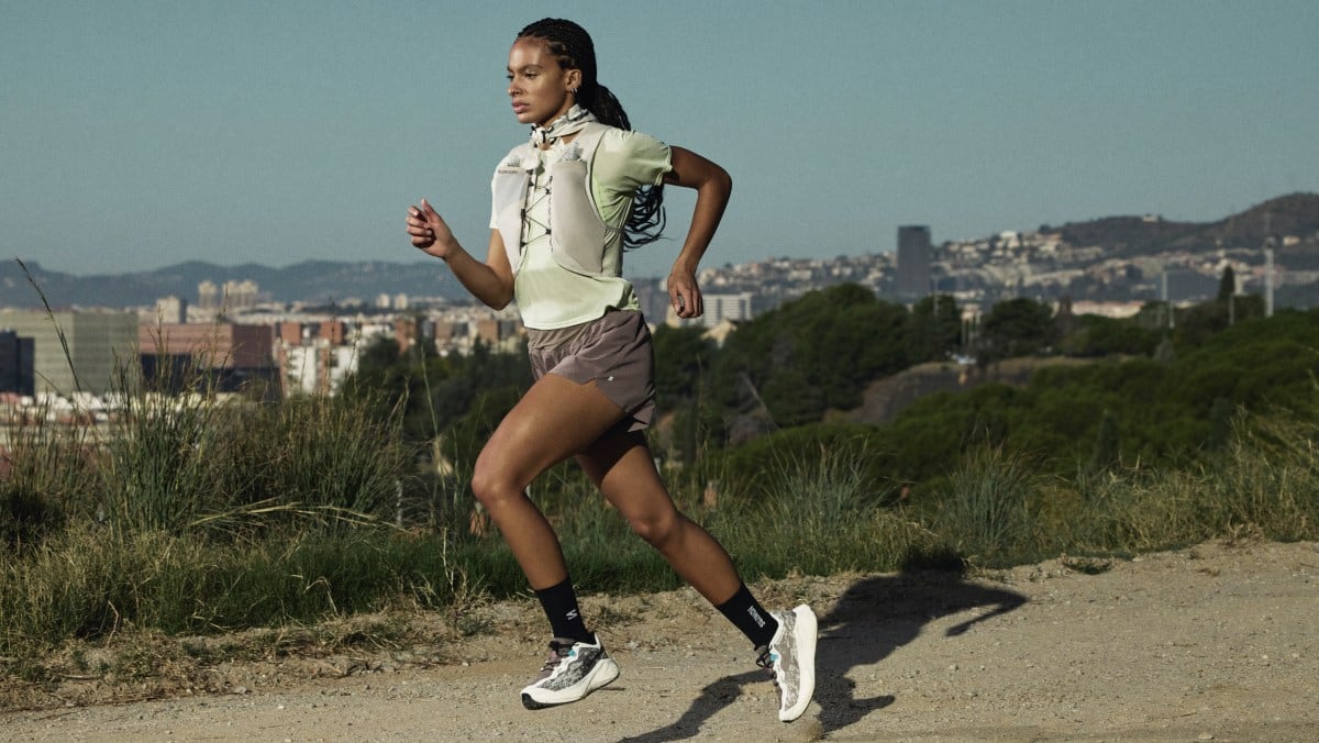 A Black woman with braids in a green top, gray running vest, and the Salomon Aero Glide 4 GRVL shoes, running along a gravel path with a city view behind her.