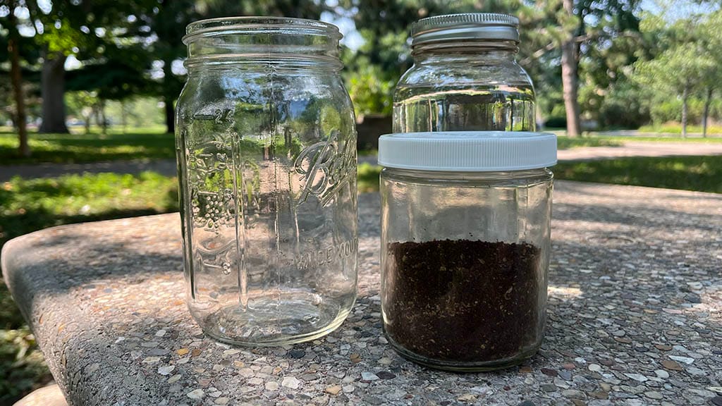 Three jars on a picnic table: One is empty and clean, one has coffee grounds in it and one has clean water in it.
