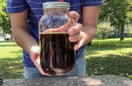 A person standing outside at a coffee table holds a jar of finished and filtered cold-brew concentrate at the camera