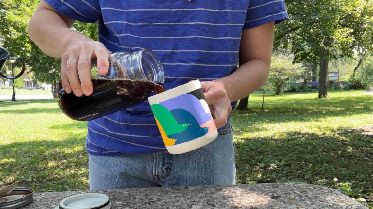 A person standing outside at a coffee table pours coffee into a travel or camp mug