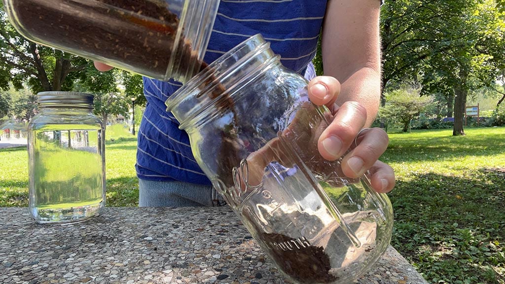 A person standing outside at a coffee table pours coffee grounds into an empty glass jar
