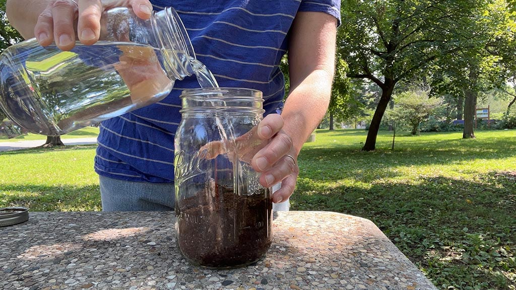 A person standing outside at a picnic table pours cold water over coffee grounds in a glass jar