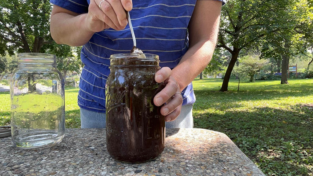A person standing outside at a coffee table stirs coffee grounds and water together in a jar.