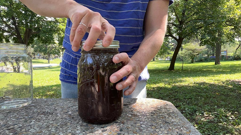 A person standing outside at a coffee table puts the lid on the glass jar full of coffee grounds and water