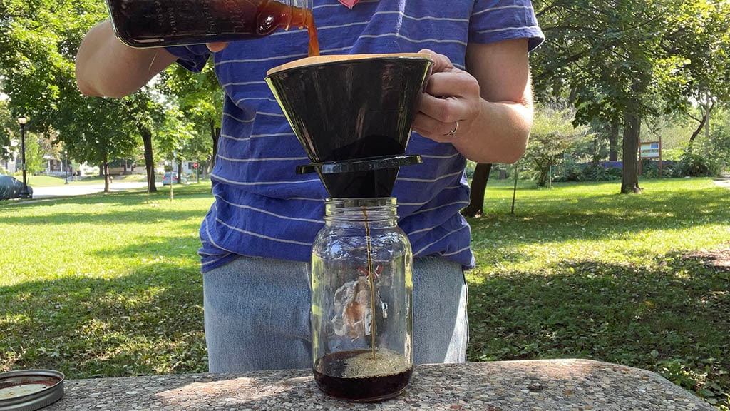 A person standing outside at a coffee table pours cold-brewed coffee into a coffee filter on top of a clean jar, as brewed coffee filters through into the bottom of the vessel