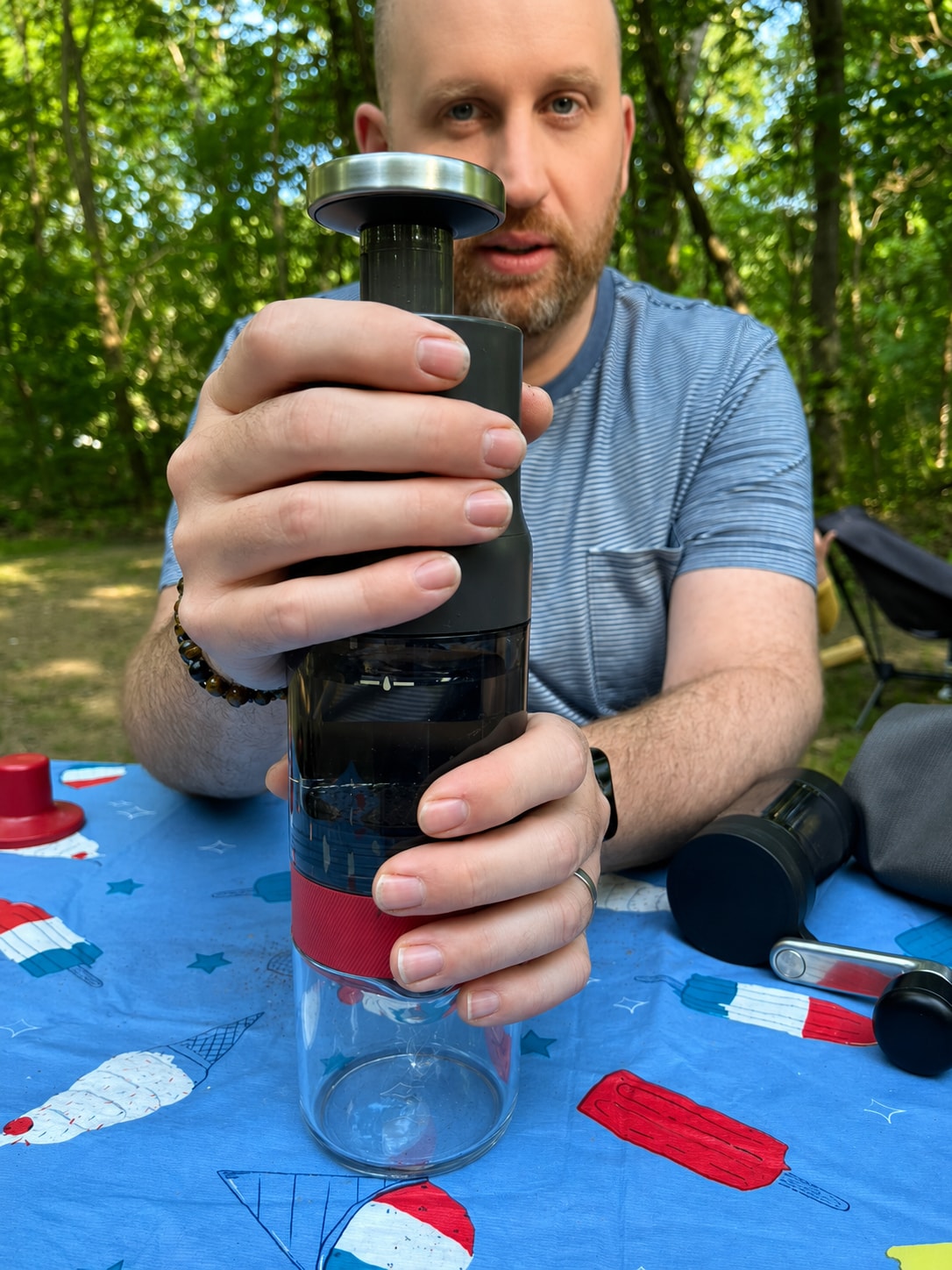 A person holds the top and bottom of an OXO coffee brewer while sitting at a picnic table in the woods.