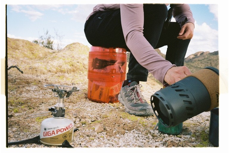 A person sitting on a bear canister, pouring coffee from a French press at a campground