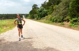 A woman in a running hydration vest and running apparel on a gravel road, walking with her hands on her hips and looking tired.