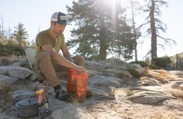A man sitting on a backpacking chair in a desert campsite opens a bear canister.