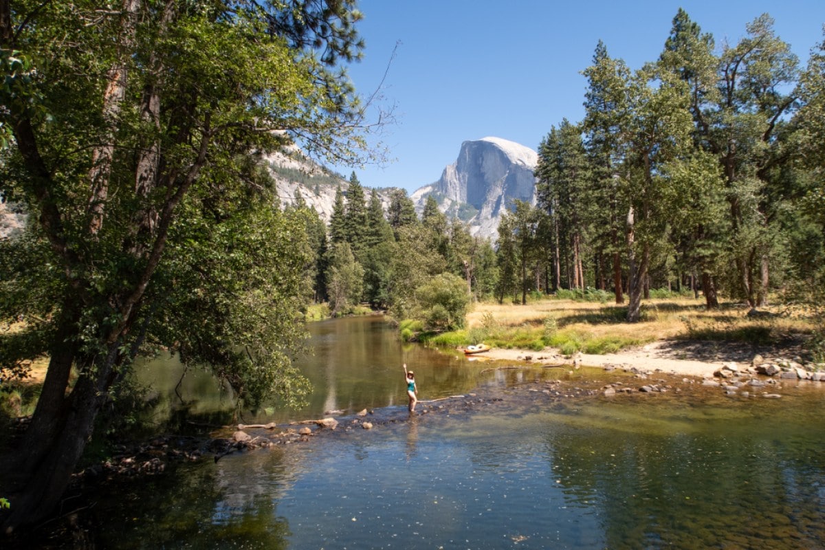 A person is shown in the middle ground waving at the camera as she crosses a body of water. Mountains are seen in the background.