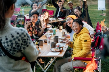 A group of people of color sit around a long camping table, cheering at a person whose back is to the camera